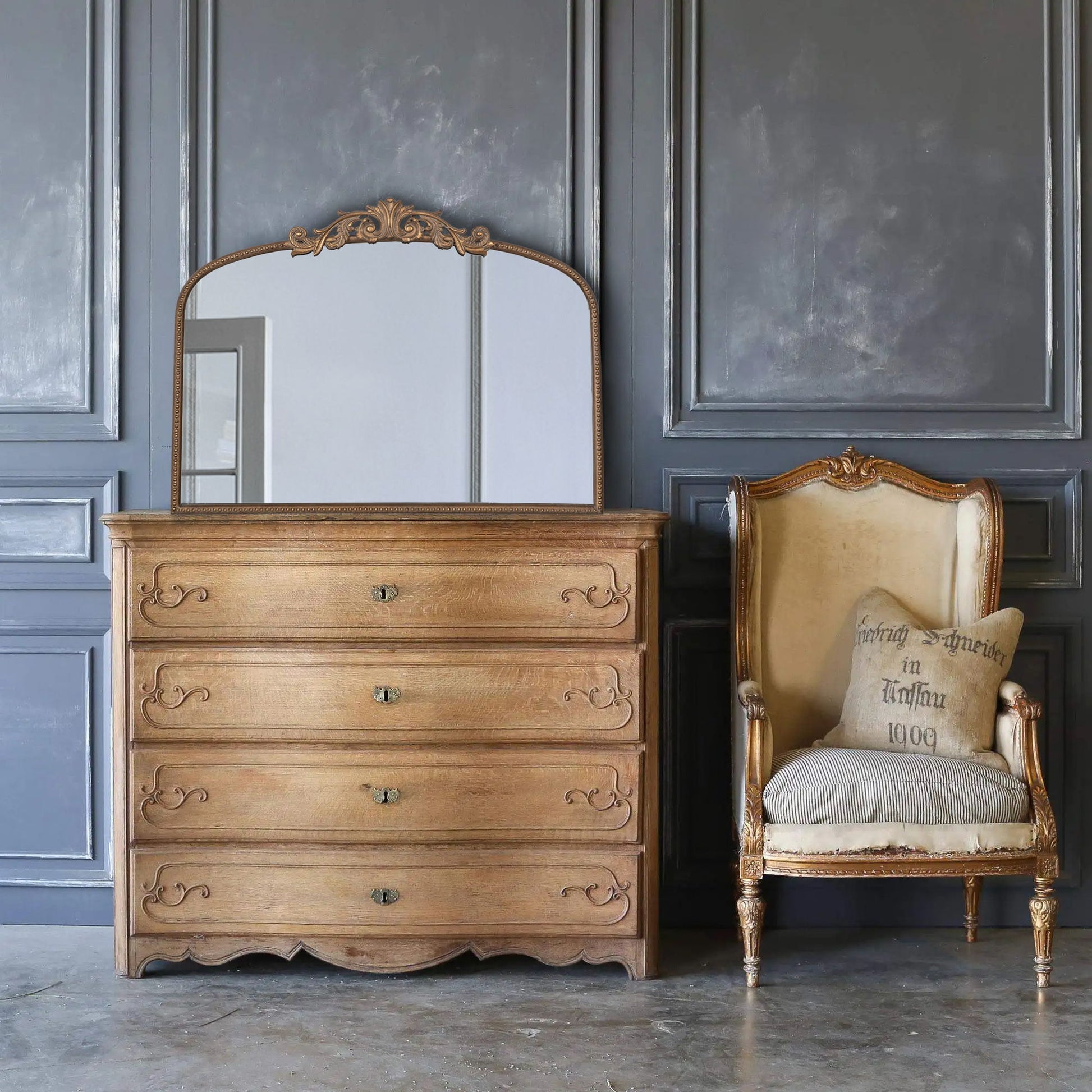 An Arch Mirror, baroque-inspired wall decor, is mounted near an antique armchair with a beige cushion and pillow, set against gray paneled walls and a polished concrete floor.