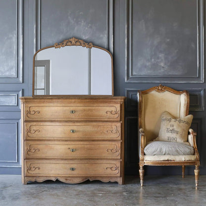 An Arch Mirror, baroque-inspired wall decor, is mounted near an antique armchair with a beige cushion and pillow, set against gray paneled walls and a polished concrete floor.
