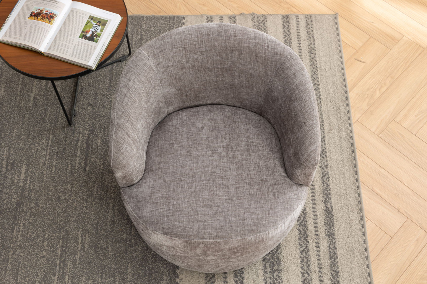 A top-down view of the Chenille Fabric Swivel Accent Armchair Barrel Chair with a gray, rounded design on a patterned rug, beside a small round table holding an open book. The flooring is light wood in a herringbone pattern.