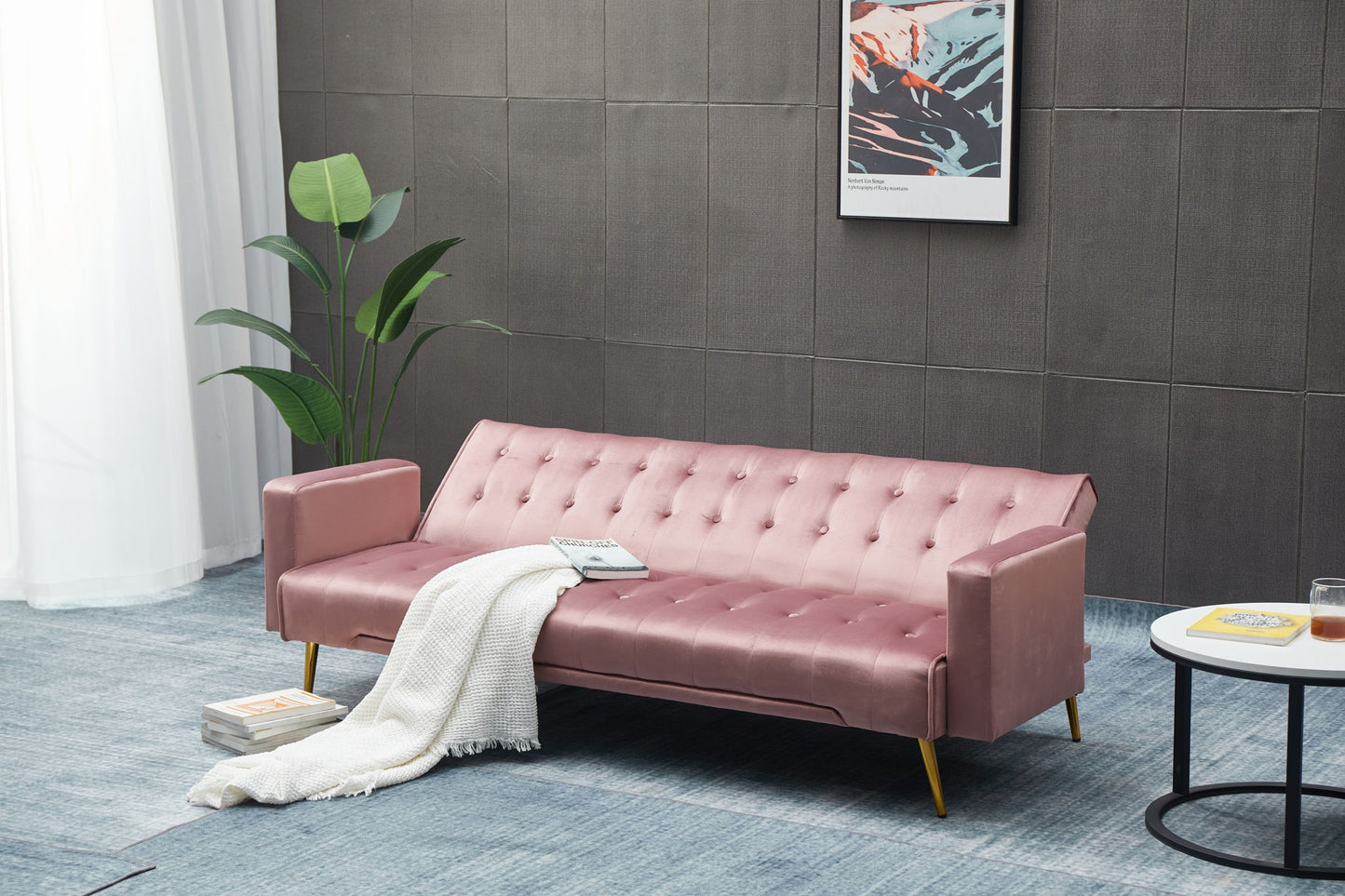 A modern living room featuring the Comfort Compact Design Sofa Bed in pink velvet, adorned with a white blanket, a potted plant, framed abstract wall art, and a round coffee table holding a drink and book.
