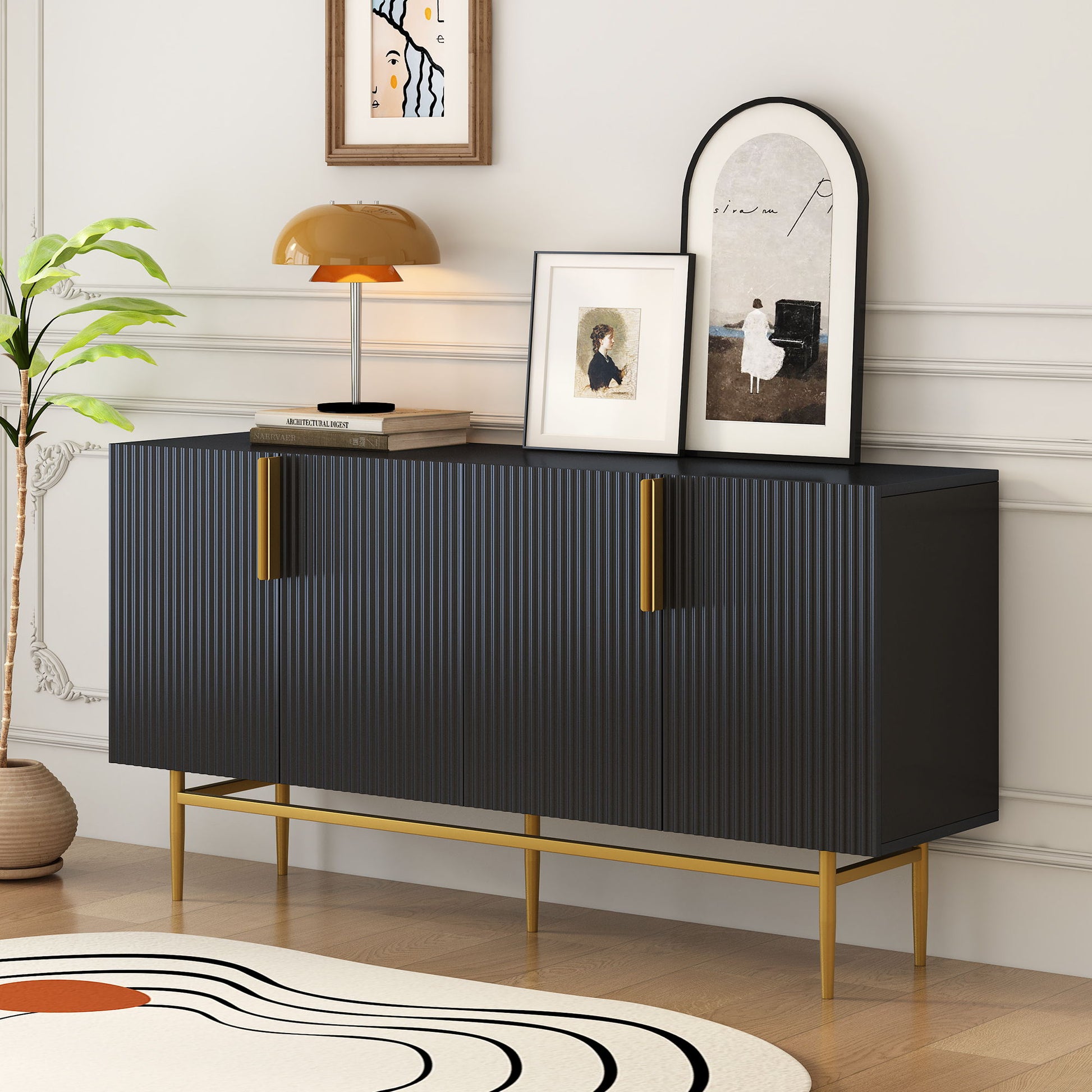 The Modern Elegant 4 Door Sideboard with gold metal handles is accented by framed art, a gold lamp, and a book on top. A potted plant and colorful rug sit beside it, all set against a white paneled wall.