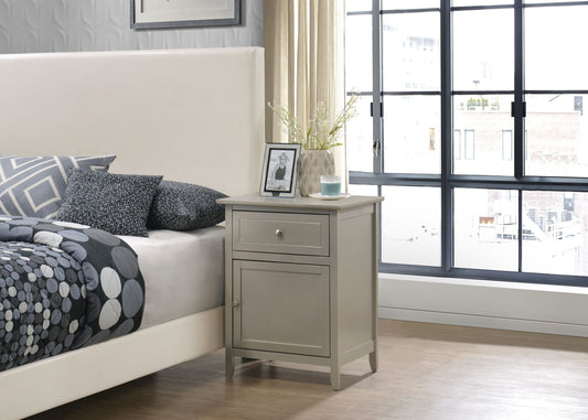 A modern bedroom with a beige bed, patterned pillows, and an Izzy - 1 Drawer /1 Door Nightstand holding a framed photo, vase with flowers, and candle beside a large window with city views and cream curtains.