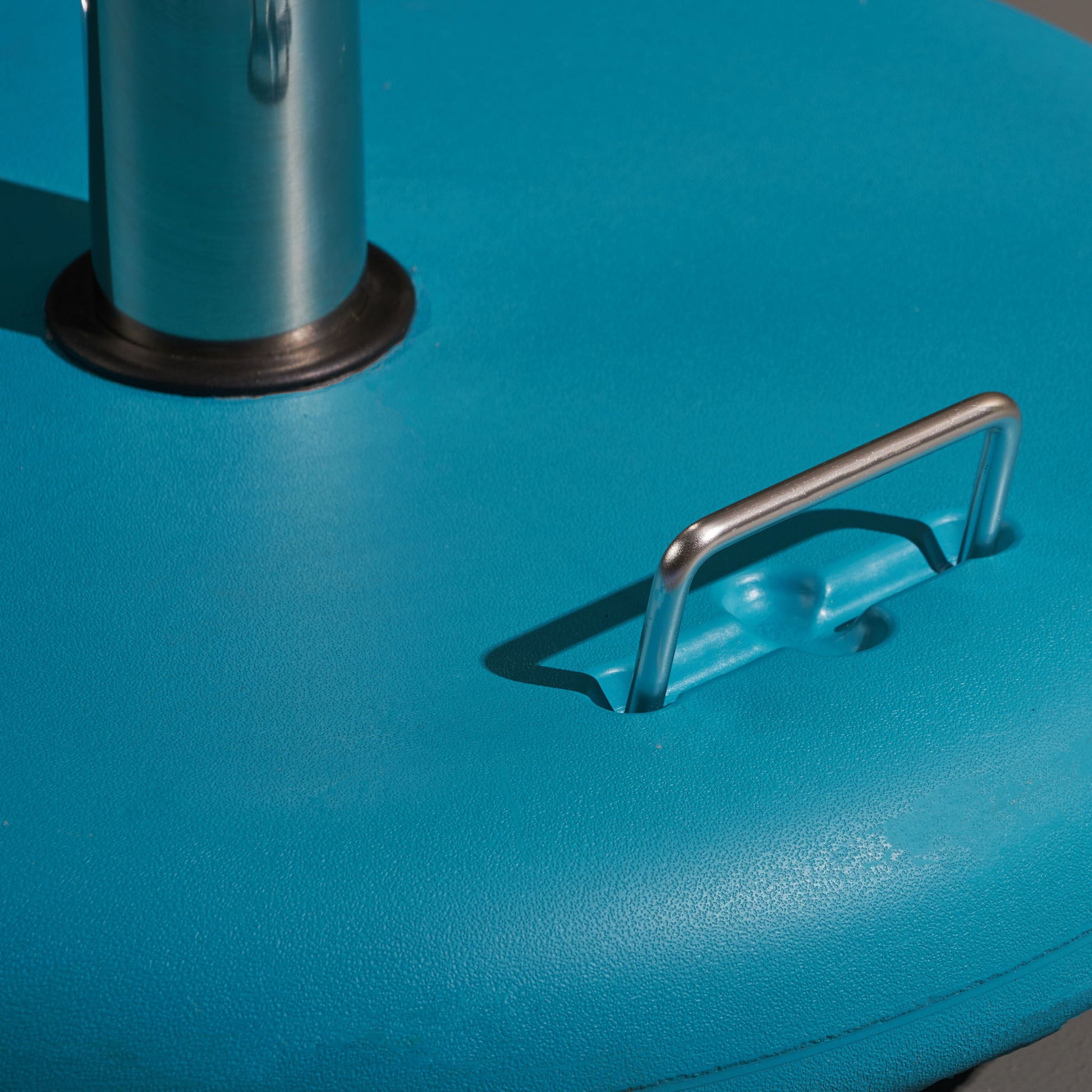 Close-up of the Wheelie - Round Umbrella Base With Stainless Steel Holder, featuring a metal handle set in the bright blue base beside a cylindrical stainless steel pole with black base, under lighting that highlights its texture and shadows.