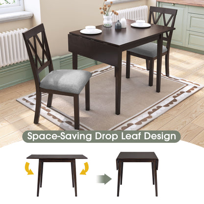 The 3 Piece Kitchen Dining Set features a drop leaf dining table and 2 upholstered chairs on a rug in a kitchen. The table is shown extended and folded, with "Space-Saving Drop Leaf Design" below, plus a vase and books on top.