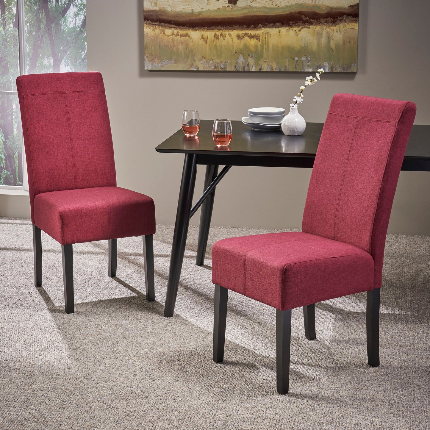 Two Pertica Dining Upholstered Armless Chairs (set of 2) with dark wooden legs sit on a beige carpet beside a black dining table holding two glasses with pink drinks and a white vase of flowers. Art decorates the wall behind.