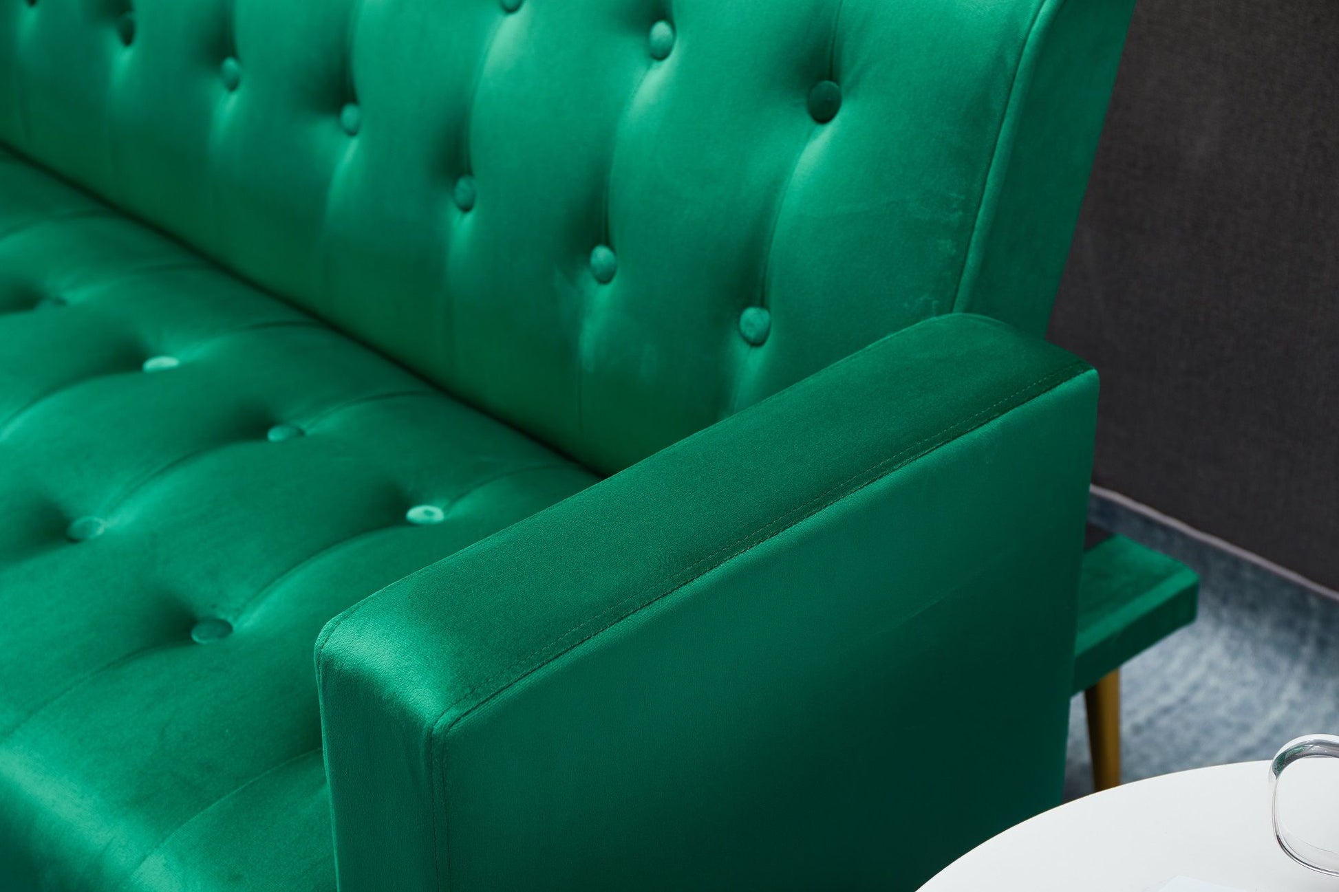 Close-up of a bright green Comfort Compact Design Sofa Bed with velvet, button-tufted upholstery and visible armrest. A white round table appears in the lower right. The background features a dark wall and gray floor.