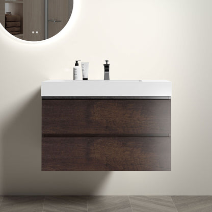 The Alice wall-mounted bathroom vanity features a modern design with a white one-piece sink basin, two dark wood drawers for storage, and a round mirror above. Two bottles rest on the counter against neutral light-colored walls and floor.