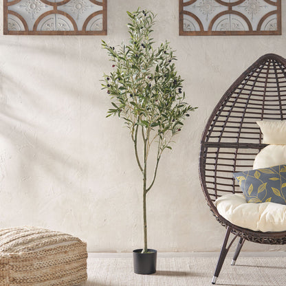 An Artificial Olive Tree stands indoors beside a wicker egg chair with cushions and a patterned pouf, set against a light textured wall with decorative panels.