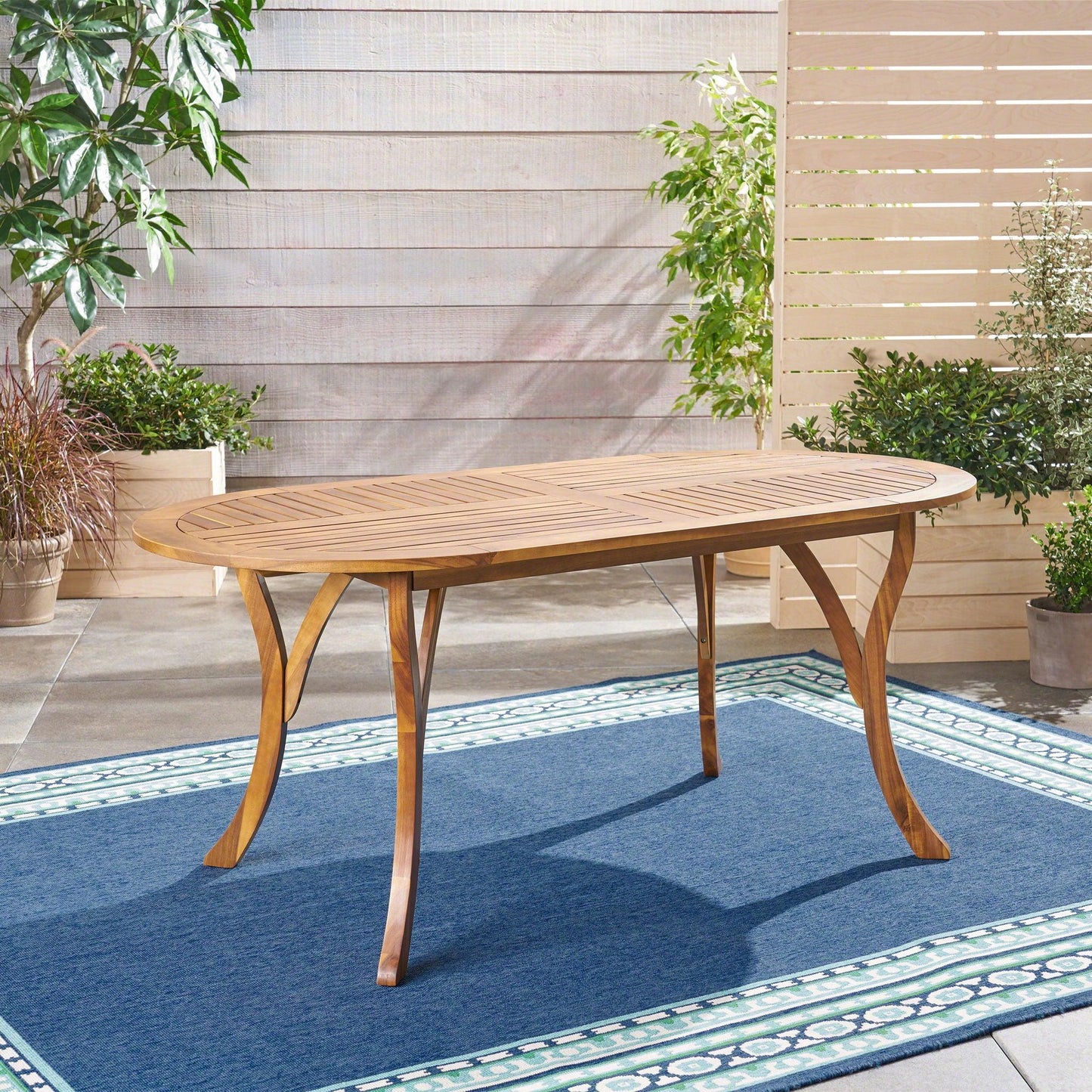 An Outdoor Table With Smooth Lines, Slat Top stands on a blue outdoor rug in a patio, surrounded by potted plants and greenery against a wooden slat fence.