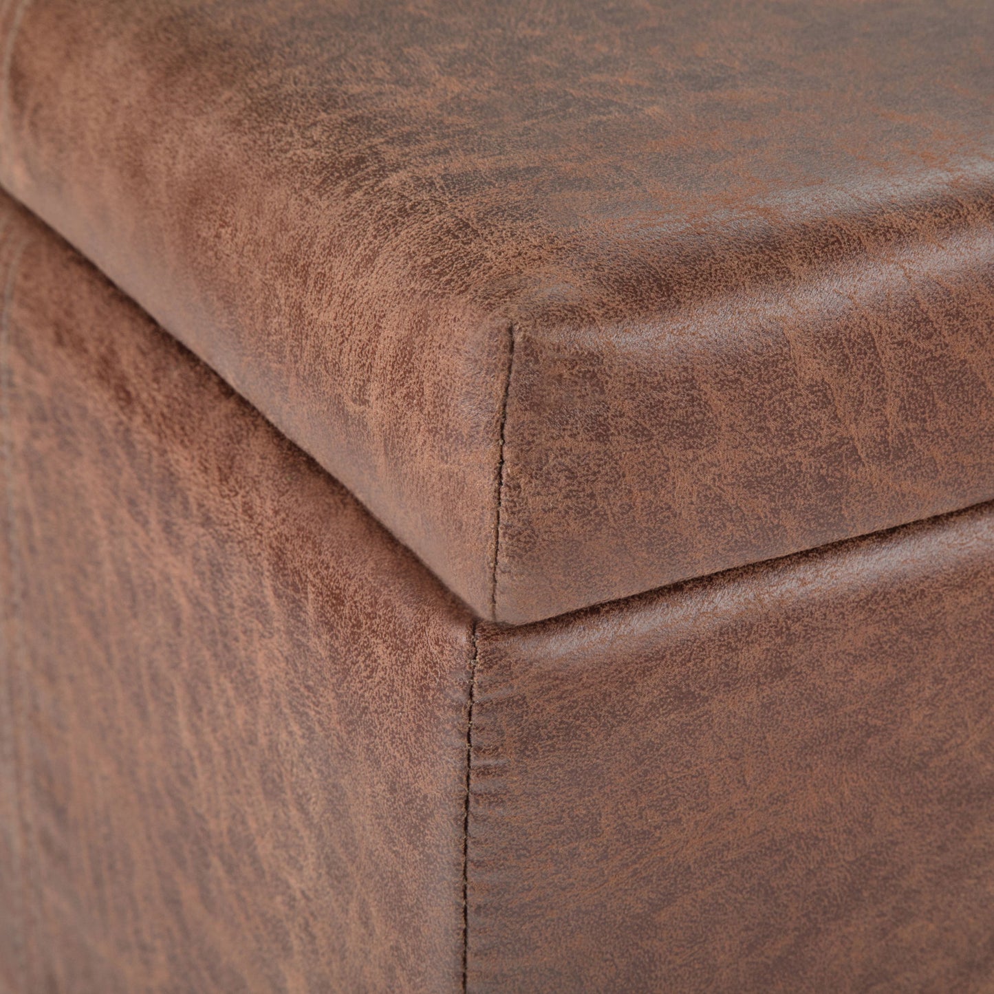 Close-up of the corner of the Dover - Upholstered Storage Ottoman Bench, highlighting its brown faux leather texture and precise stitching along the edge.
