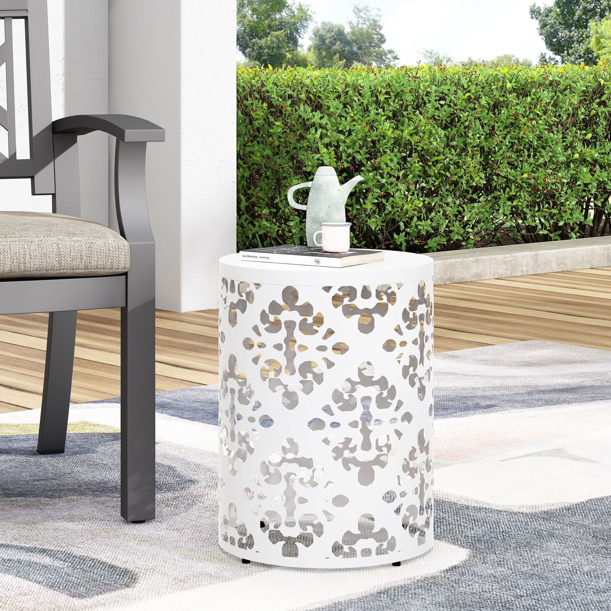 The Outdoor Metal Modern Design End Table, featuring a white round shape and decorative cutout pattern, sits on a rug beside a cushioned outdoor chair. A light green teapot and cup are placed on top, with hedges and trees in the background.