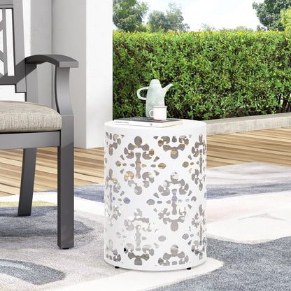 The Outdoor Metal Modern Design End Table, featuring a white round shape and decorative cutout pattern, sits on a rug beside a cushioned outdoor chair. A light green teapot and cup are placed on top, with hedges and trees in the background.