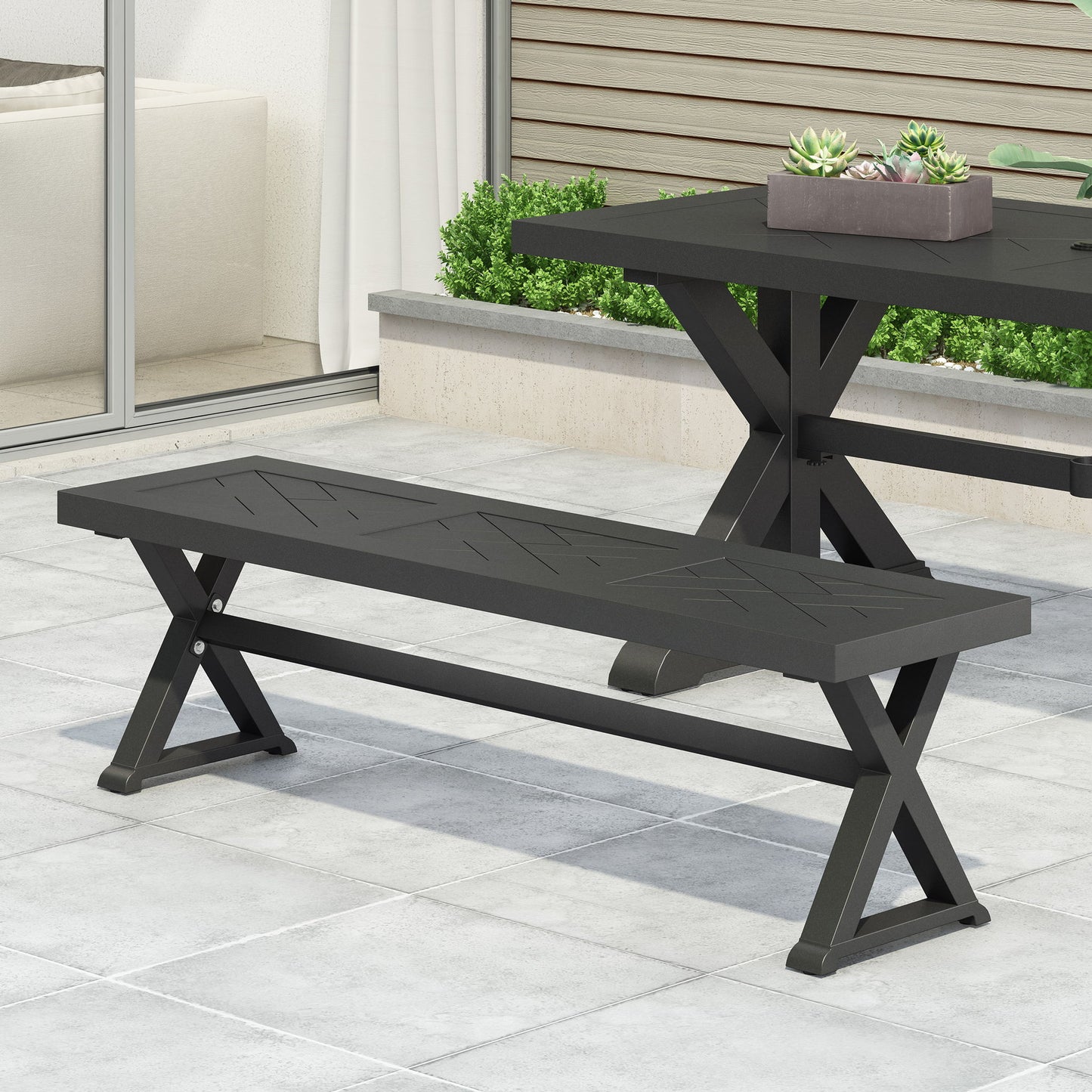 A Modern Aluminum Outdoor Dining Bench With X Base in black sits on a tiled patio beside a matching table with a rectangular planter of succulents, framed by glass doors and a small garden bed.