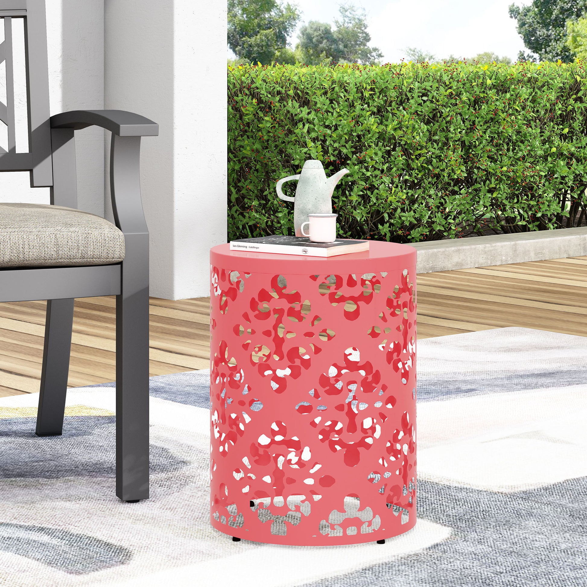 The Outdoor Metal Modern Design End Table, featuring a red cylindrical shape with decorative cutouts, stands on a patio rug beside a gray chair. A white teapot and cup are on top, with green bushes and trees in the background.