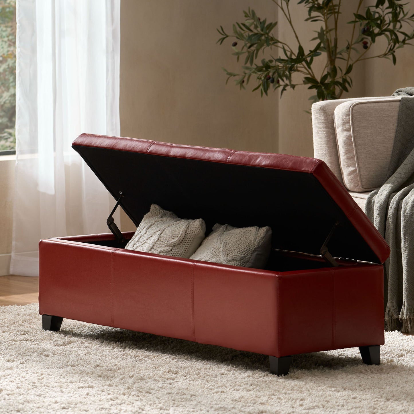 The Glouster - Contemporary Storage Ottoman, shown open in a cozy, sunlit living room, reveals two gray knit pillows inside. It sits on a beige rug beside a sofa and a plant.