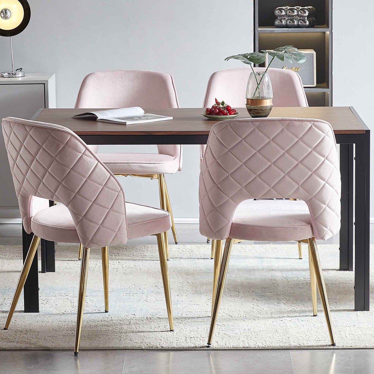 A modern dining area features a wooden table with four Velvet Dining Chairs With Metal Legs And Hollow Back Upholstered Dining Chairs in pink, an open book, a bowl of cherries, and a glass vase with a plant. Shelves and decor appear in the background.