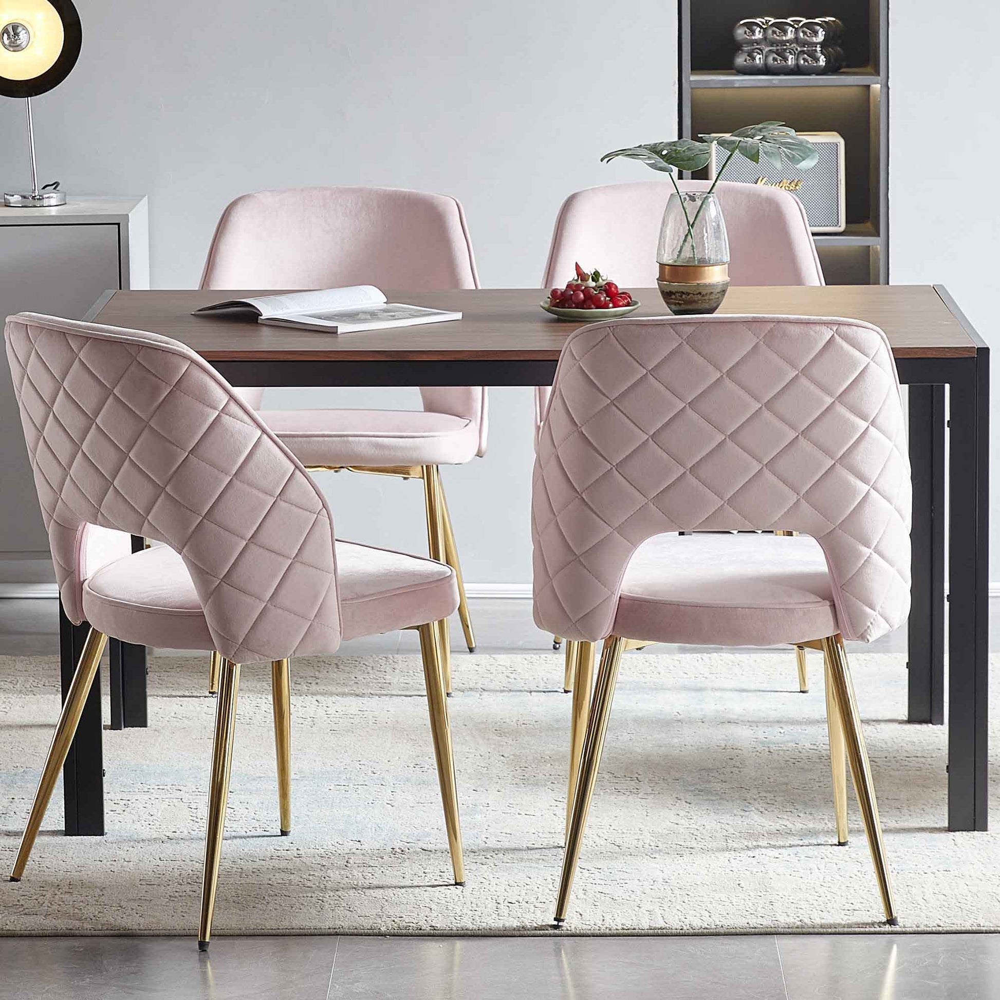 A modern dining area features a wooden table with four Velvet Dining Chairs With Metal Legs And Hollow Back Upholstered Dining Chairs in pink, an open book, a bowl of cherries, and a glass vase with a plant. Shelves and decor appear in the background.