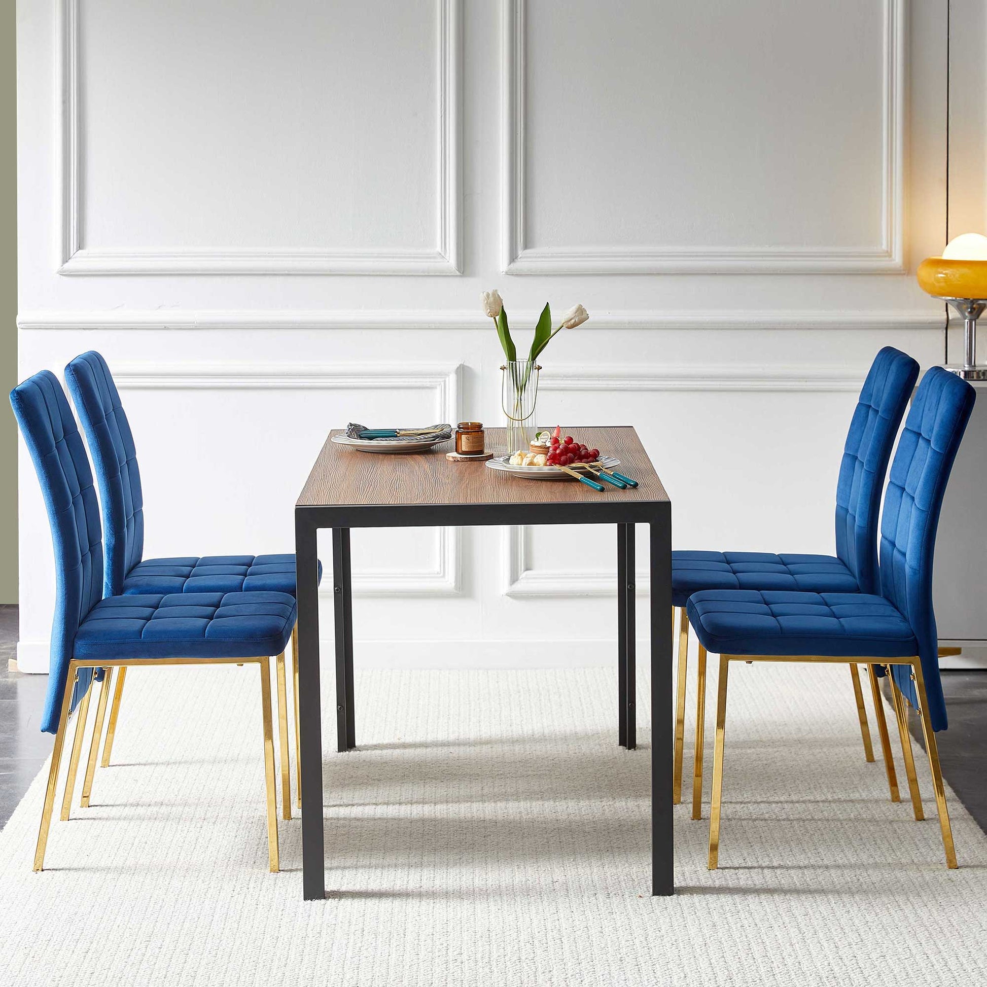 A modern dining area features the 5 Pieces Dining Set, including a creative table and four velvet high back Nordic chairs with golden legs, plus a white vase, plates, and grapes atop a light rug against a white paneled wall.