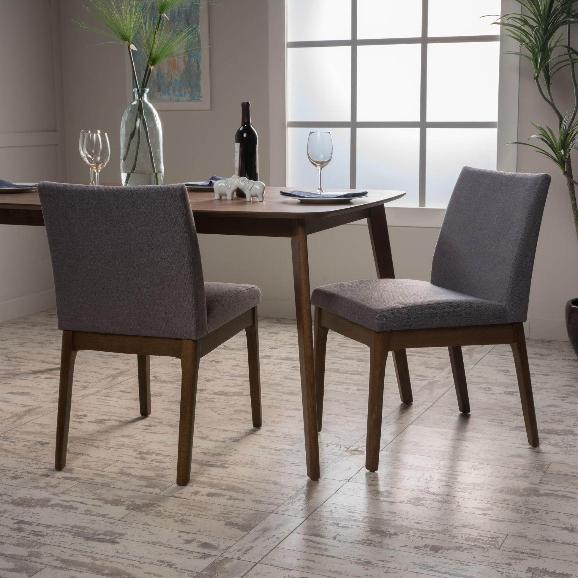 Two Contemporary Fabric Comfort Dining Chairs (Set of 2) with gray cushions are placed by a wooden dining table set with wine glasses, plates, and a wine bottle in a bright room filled with plants and featuring a frosted window.