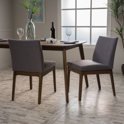Two Contemporary Fabric Comfort Dining Chairs (Set of 2) with gray cushions are placed by a wooden dining table set with wine glasses, plates, and a wine bottle in a bright room filled with plants and featuring a frosted window.