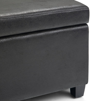 Close-up of the corner of the Avalon - Storage Ottoman Bench in distressed black faux leather, featuring visible stitching and short black legs that highlight its texture and material.
