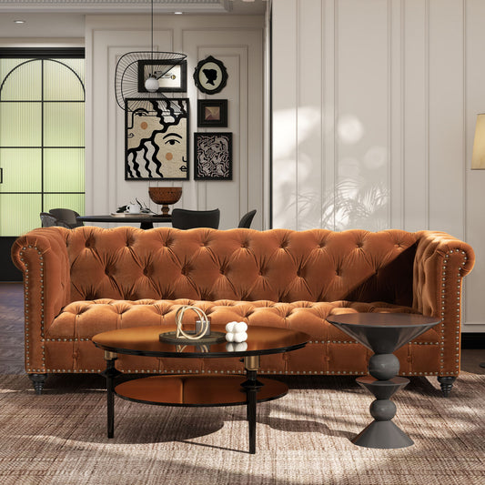 A modern living room with the Alto Tufted Chesterfield Sofa in brown velvet, two round coffee tables, a patterned rug, and abstract art. The background features a dining area and large arched windows.