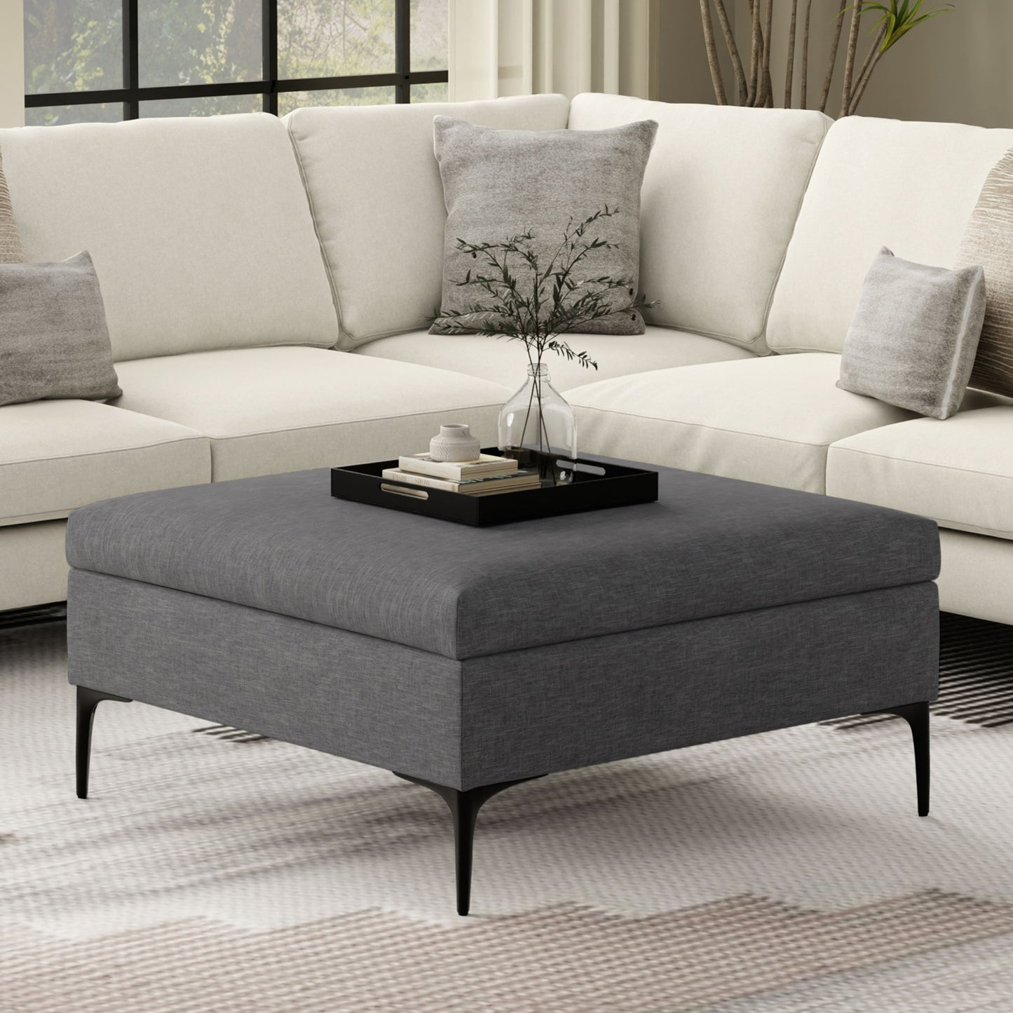 A modern living room with a white sectional sofa and the Rebecca multifunctional coffee table storage ottoman with lift up lid, styled with a black tray holding a plant, books, and a cup. Large windows and neutral-toned decor complete the space.