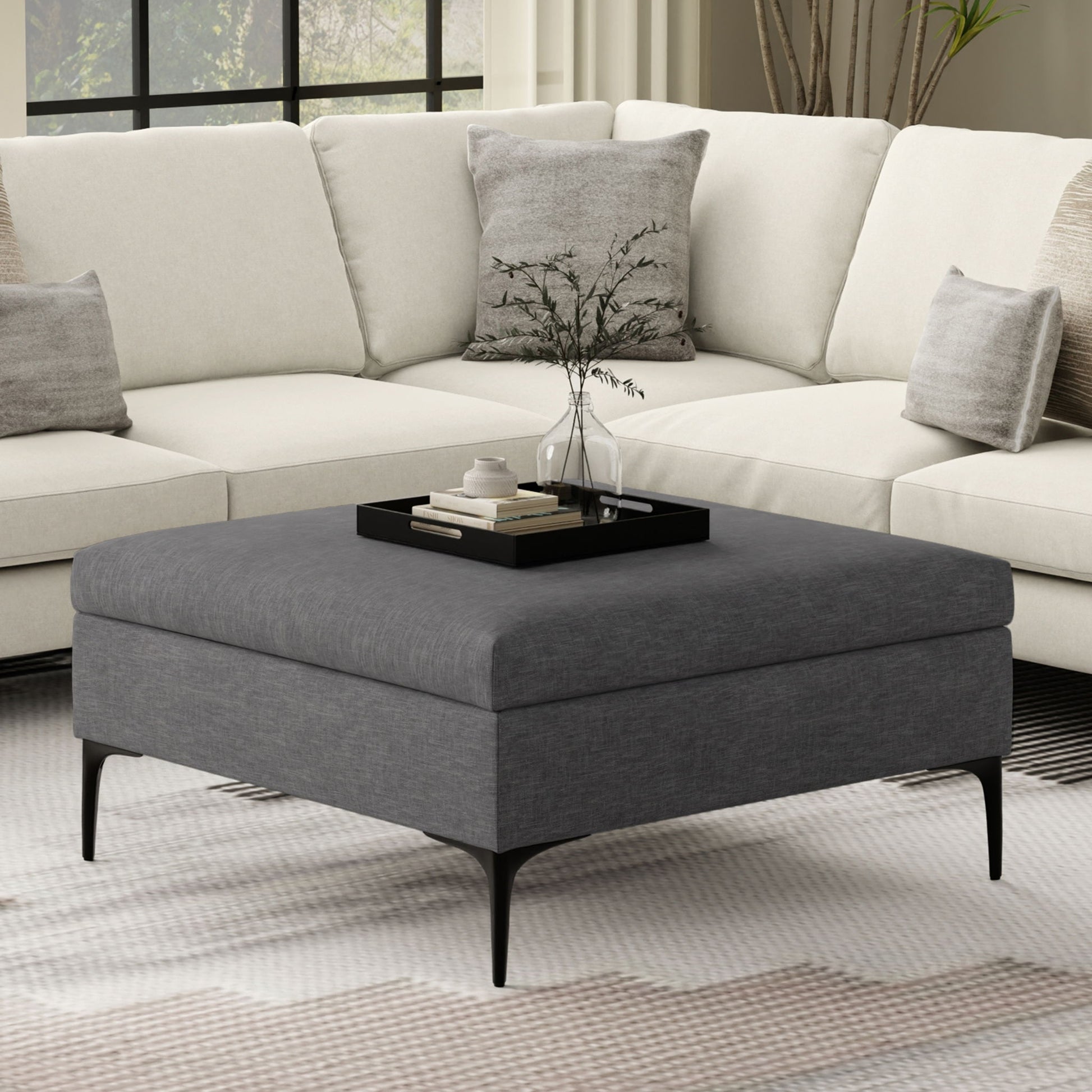 A modern living room with a white sectional sofa and the Rebecca multifunctional coffee table storage ottoman with lift up lid, styled with a black tray holding a plant, books, and a cup. Large windows and neutral-toned decor complete the space.