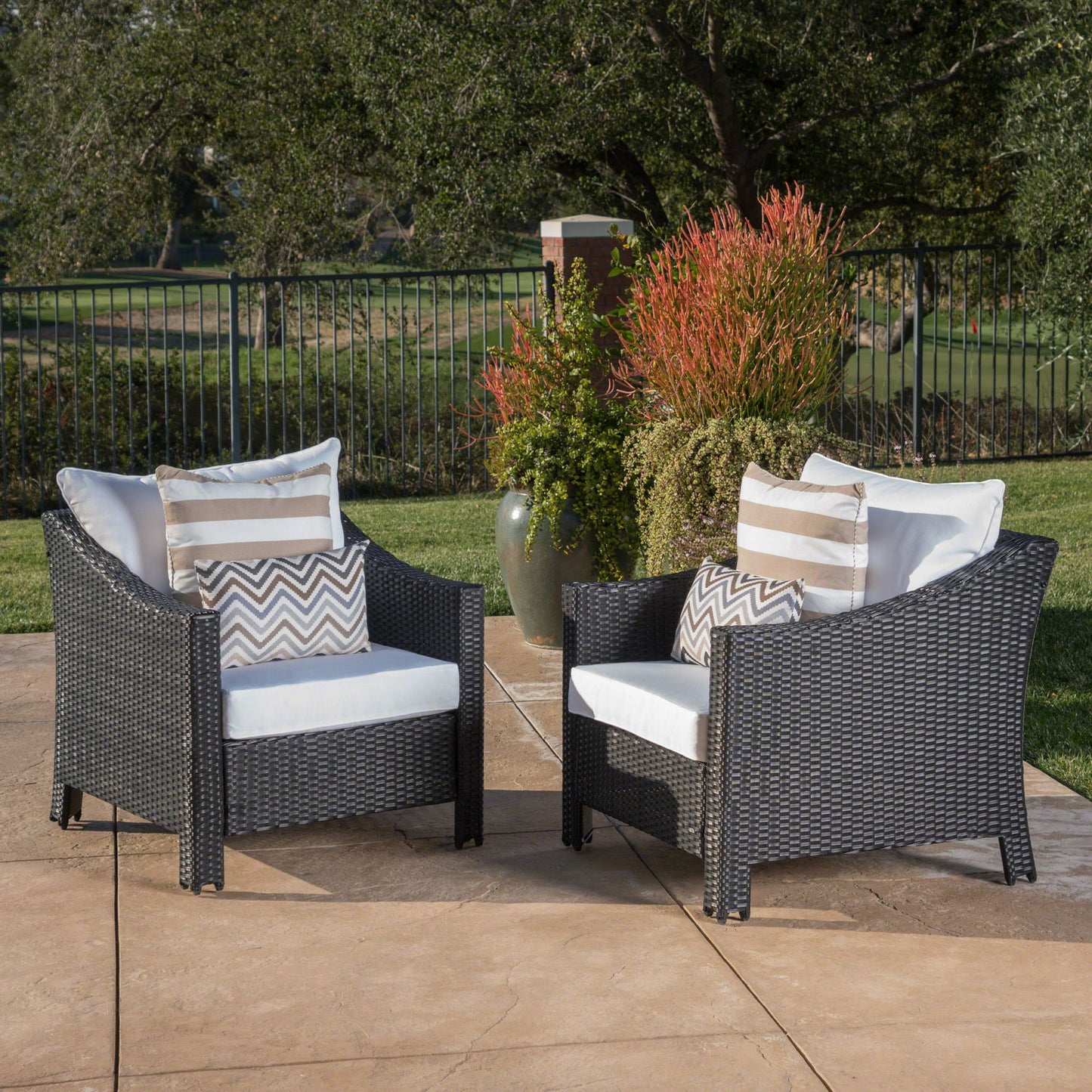 Two Antibes armchairs with water resistant cushions sit on a patio. Green plants and a large vase are behind the chairs, with a metal fence and trees visible in the background.