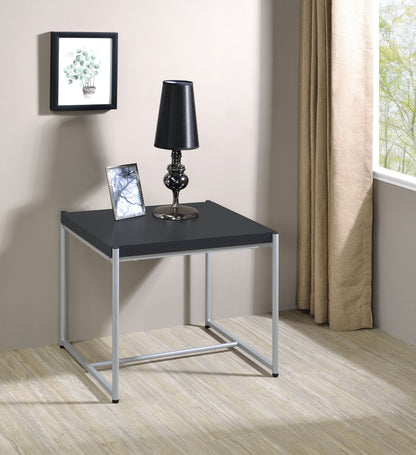 The Lennox 3 Piece Coffee and End Table Set features a compact black table with silver legs, topped with a black lamp and framed photo. Behind it are beige curtains, artwork, a large window overlooking greenery, and light wood floors.