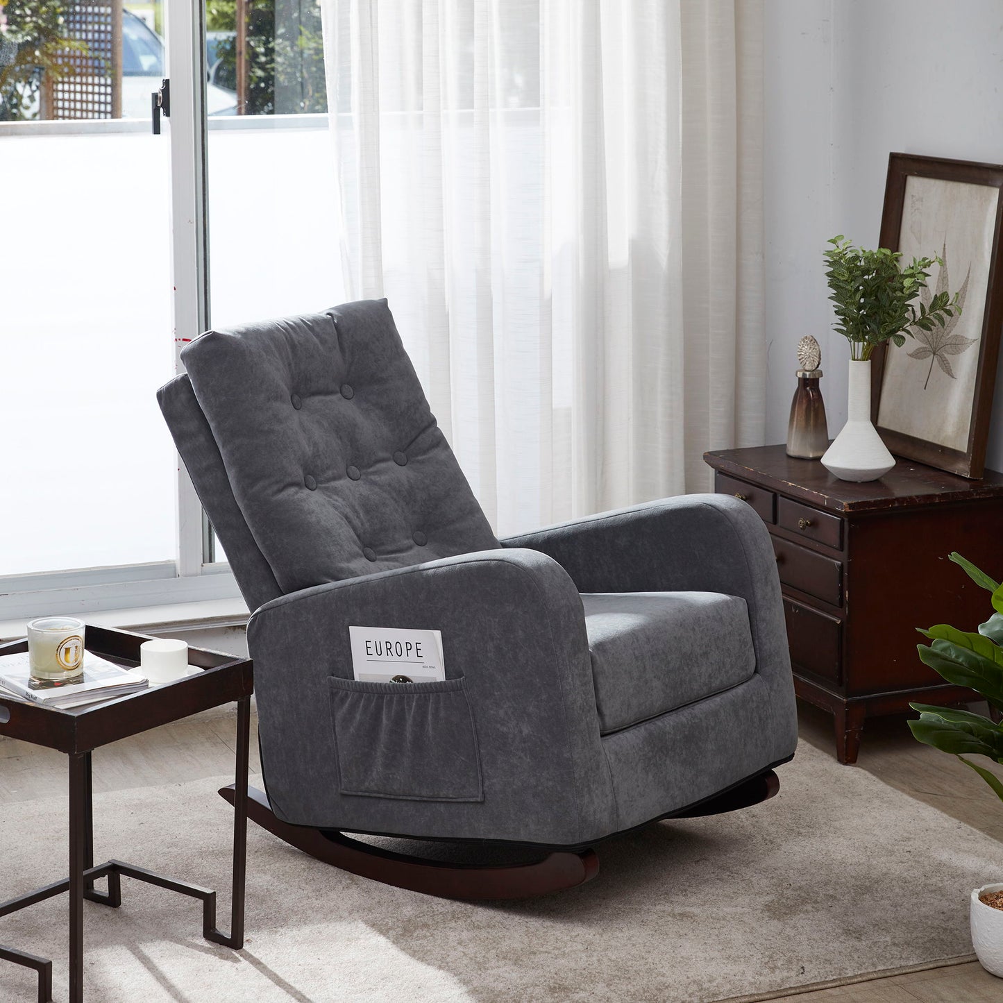 A Single Sofa Reclining Japanese Chair in gray sits on a beige rug by a window with sheer curtains, next to a wooden side table and chest of drawers. A small plant and decor accent the bright living room scene.