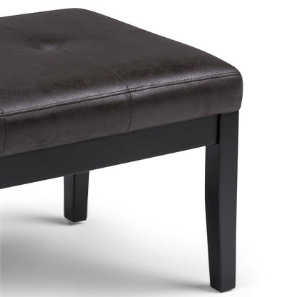 The Lacey Upholstered Tufted Ottoman Bench features a dark brown faux leather seat and black wooden legs, shown close-up against a white background.