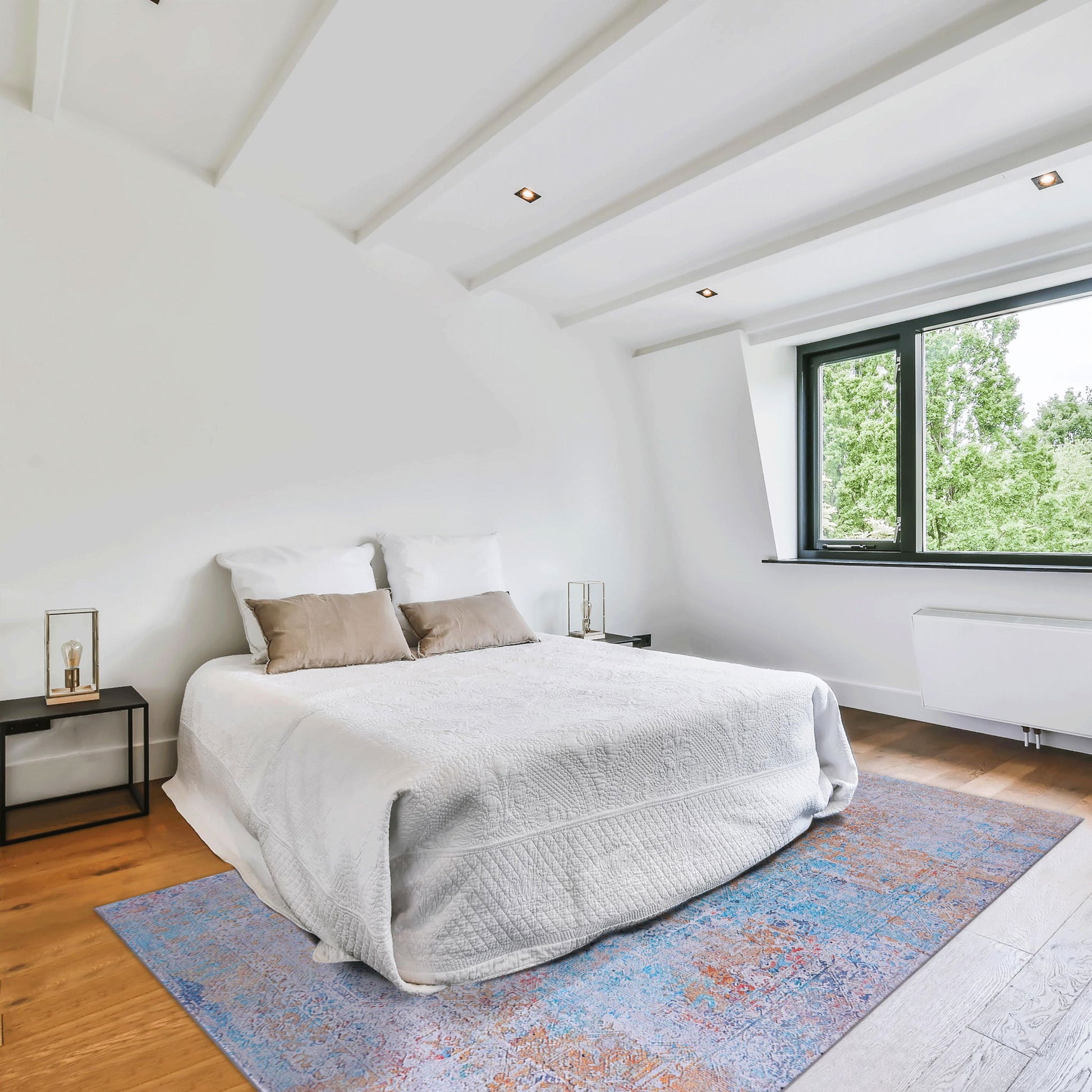 A bright, minimalist bedroom with a white bed, beige pillows, nightstands, a large window with a green view, and the Maval - Unique Washable Area Rug adding color to the wooden floor.