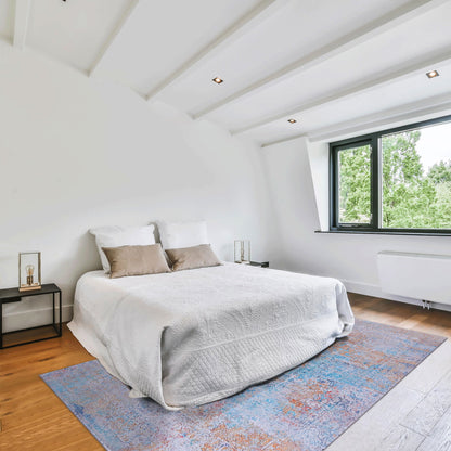 A bright, minimalist bedroom with a white bed, beige pillows, nightstands, a large window with a green view, and the Maval - Unique Washable Area Rug adding color to the wooden floor.