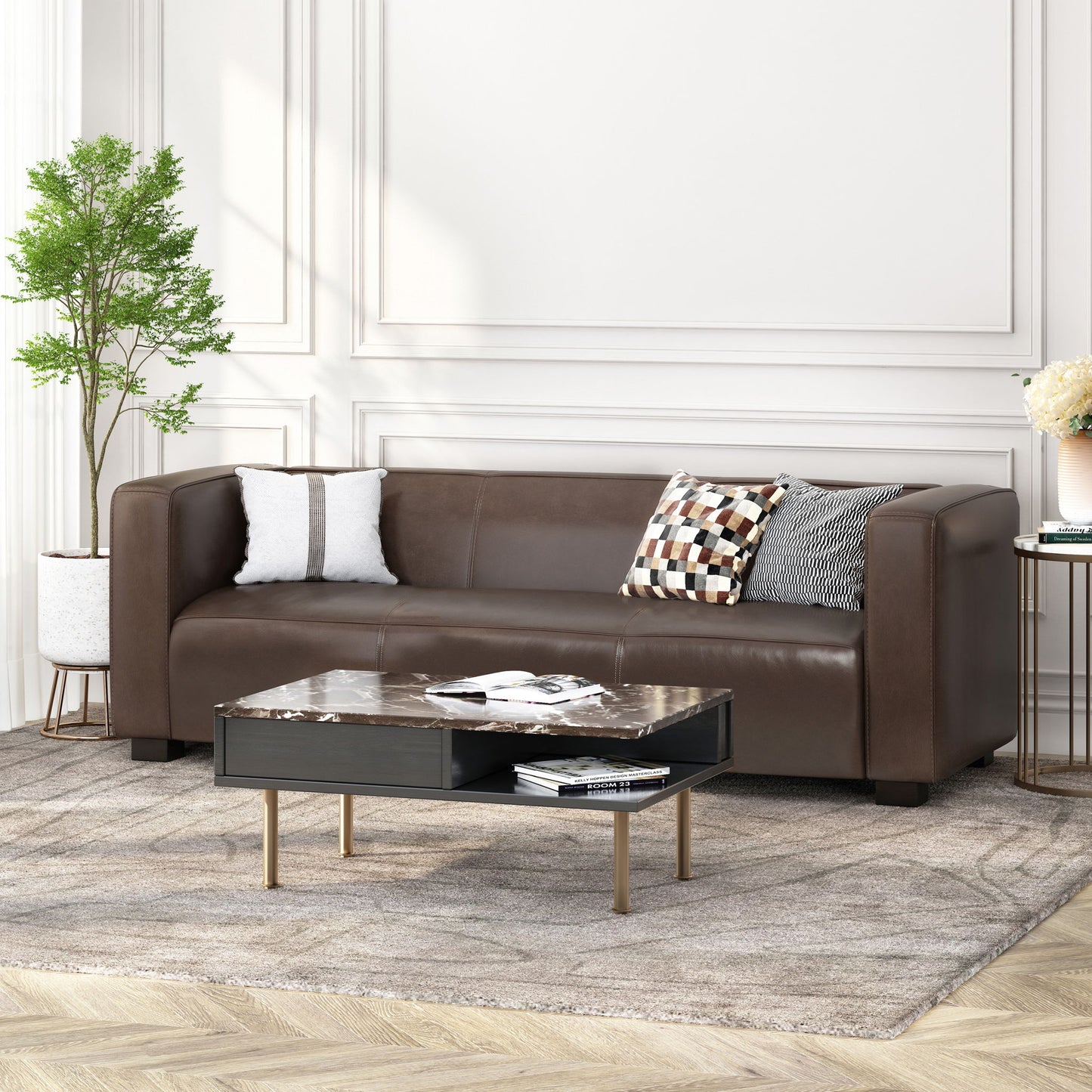 A modern living room featuring the Contemporary 3 Seater Sofa With Tuxedo Design in brown leather, three decorative pillows, a marble-top coffee table with books, a potted plant, and a light patterned rug on wooden flooring against white paneled walls.