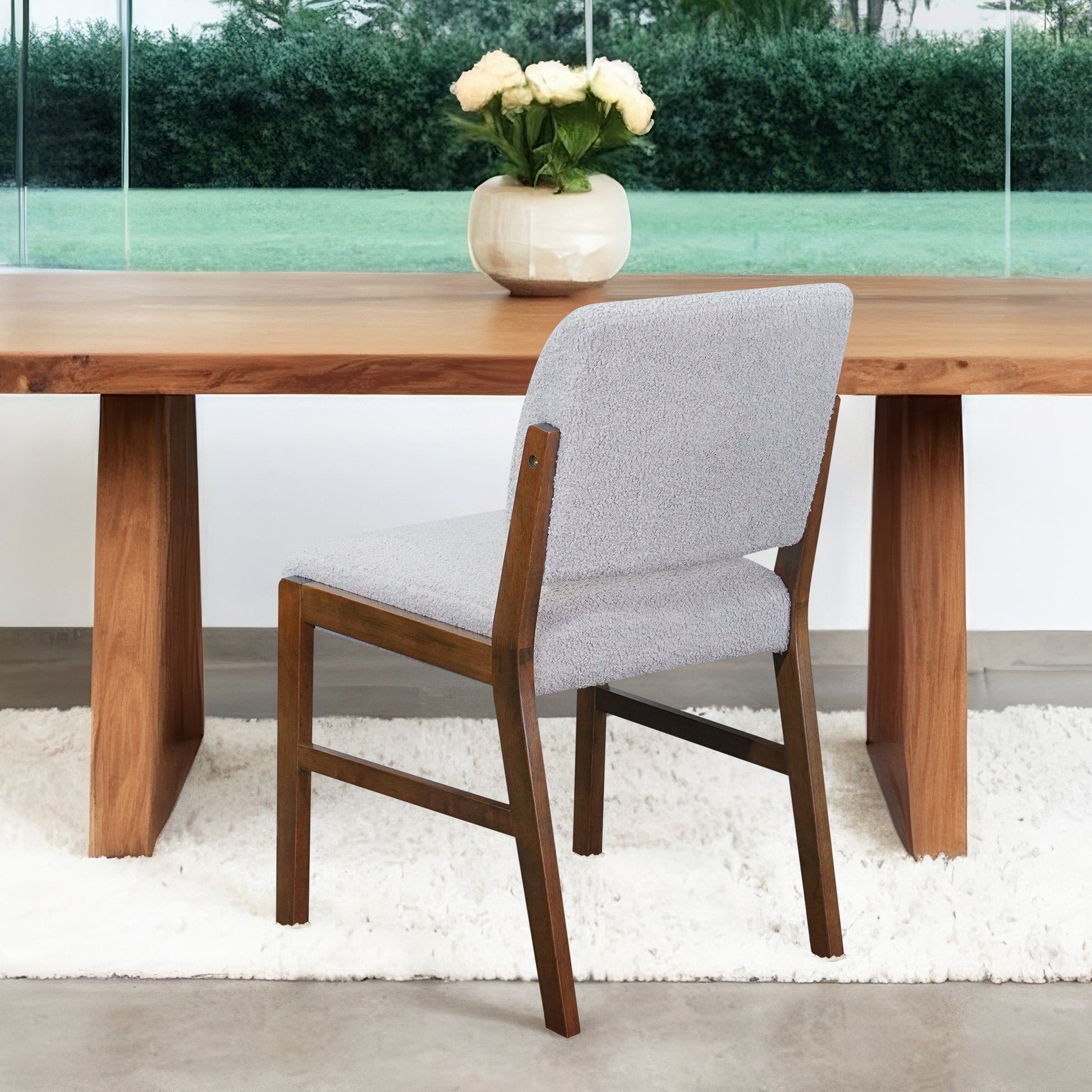 A Caldwell Dining Chair (Set of 2) in light gray sits by a wooden table with a white vase of pale roses. A window displays a grassy yard outside, and a white rug lies beneath the furniture.