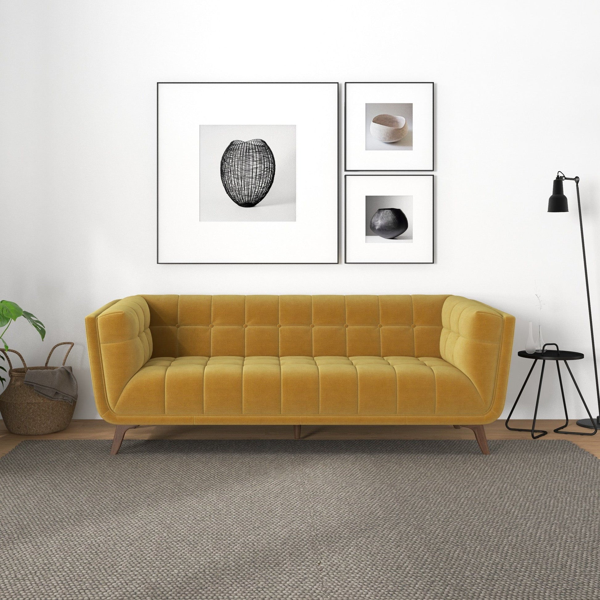 A modern living room with the Addison Mid-Century Modern Tufted Sofa in mustard yellow, a neutral rug, black floor lamp, small round side table, two baskets holding a plant, and four minimalist art prints framed on a white wall.
