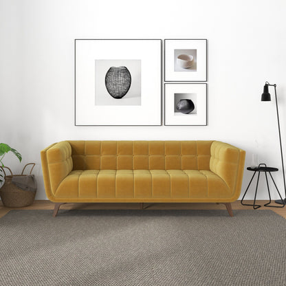 A modern living room with the Addison Mid-Century Modern Tufted Sofa in mustard yellow, a neutral rug, black floor lamp, small round side table, two baskets holding a plant, and four minimalist art prints framed on a white wall.
