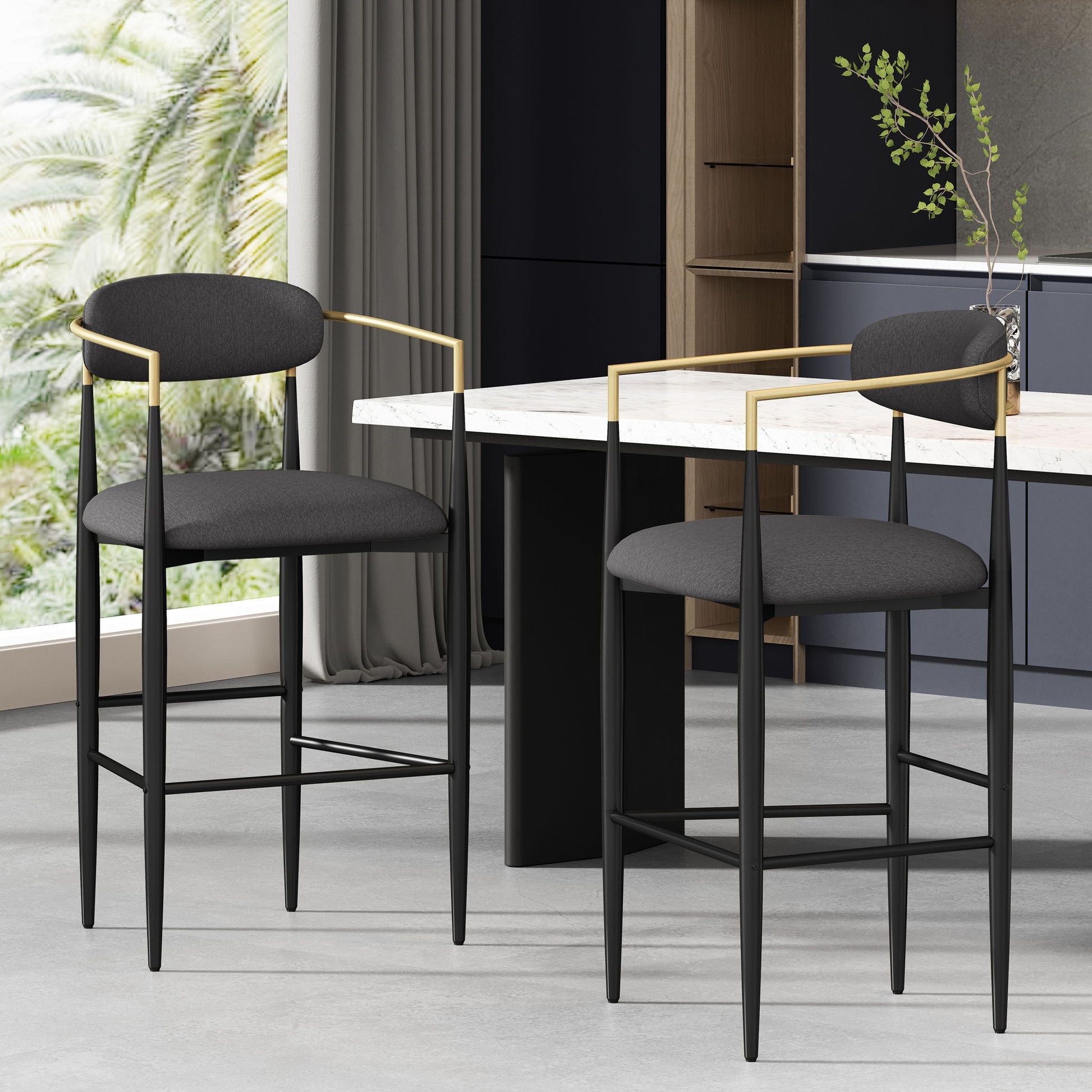 Two Counter Height Upholstered Barstools (Set of 2) with black fabric, gold-accent armrests, and rounded backs sit by a marble-topped table in a modern kitchen with large windows revealing lush green palm trees outside.