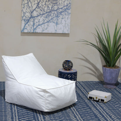 A modern room featuring the Ettie Water Resistant Fabric Bean Bag Chair in white, a blue patterned rug, small side table with a decorative sphere, potted plant, white briefcase, and framed tree branch artwork on the wall.