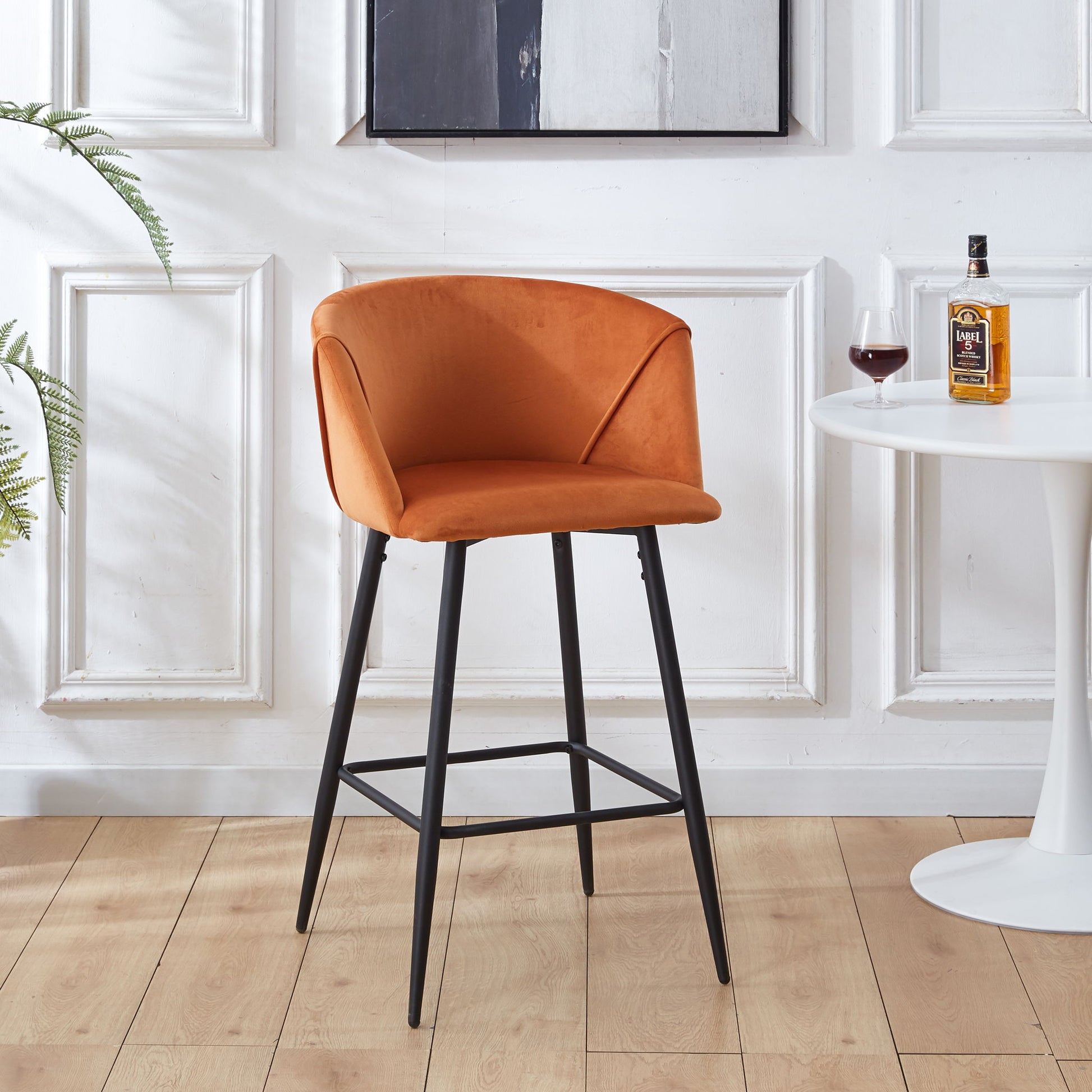 A Counter Height Bar Stool Metal Legs (Set of 2) in orange upholstery stands next to a round white table with liquor and a potted plant, set against stylish white paneled walls.
