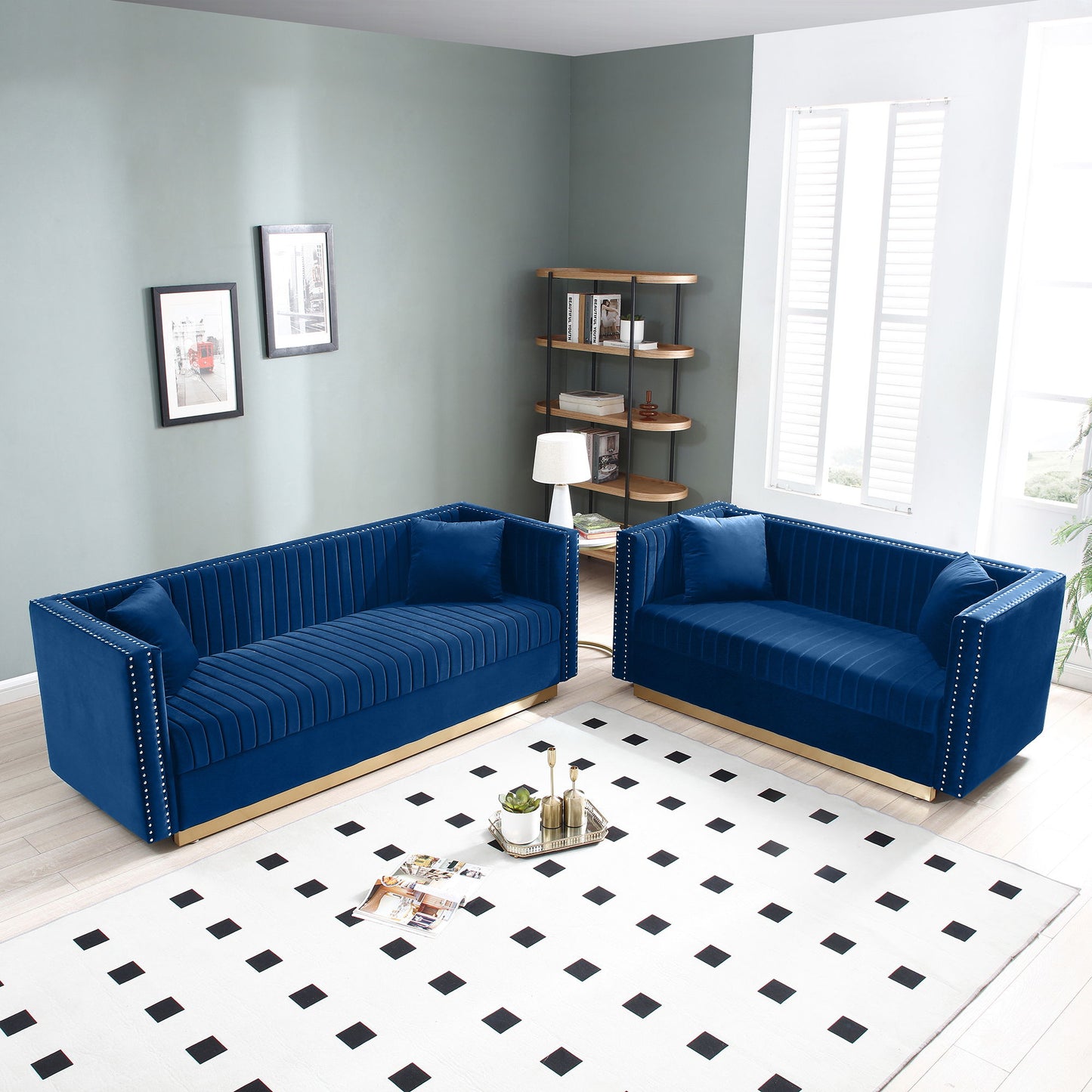 A modern living room features two Contemporary Vertical Channel Tufted Velvet Sofa Loveseats in blue, a white rug with black squares, a floor lamp, a corner shelf with books, framed art on green walls, and tall windows with white blinds.