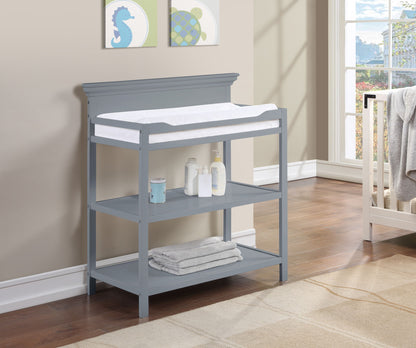 The Universal - Changing Table features a gray finish with a white pad on top, baby care essentials on the middle shelf, and folded towels below, all set in a bright nursery with wall art and a window.