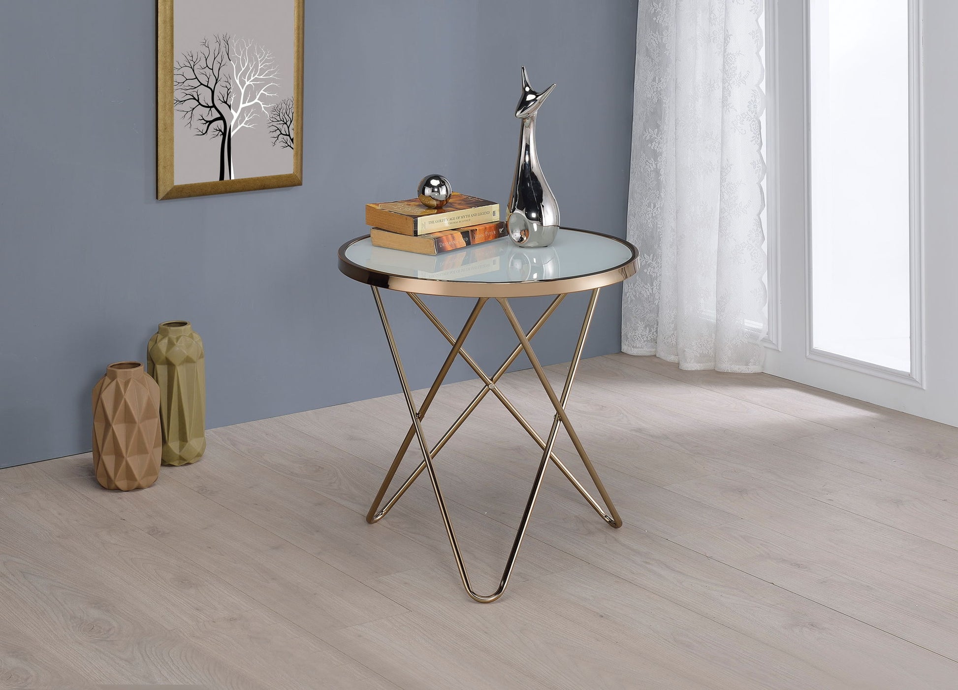 The Valora Glass Coffee Table with a gold metal frame stands on a light wood floor, displaying books and silver decor. Nearby are two vases, a framed tree print on a blue wall, and sheer curtains by the window.