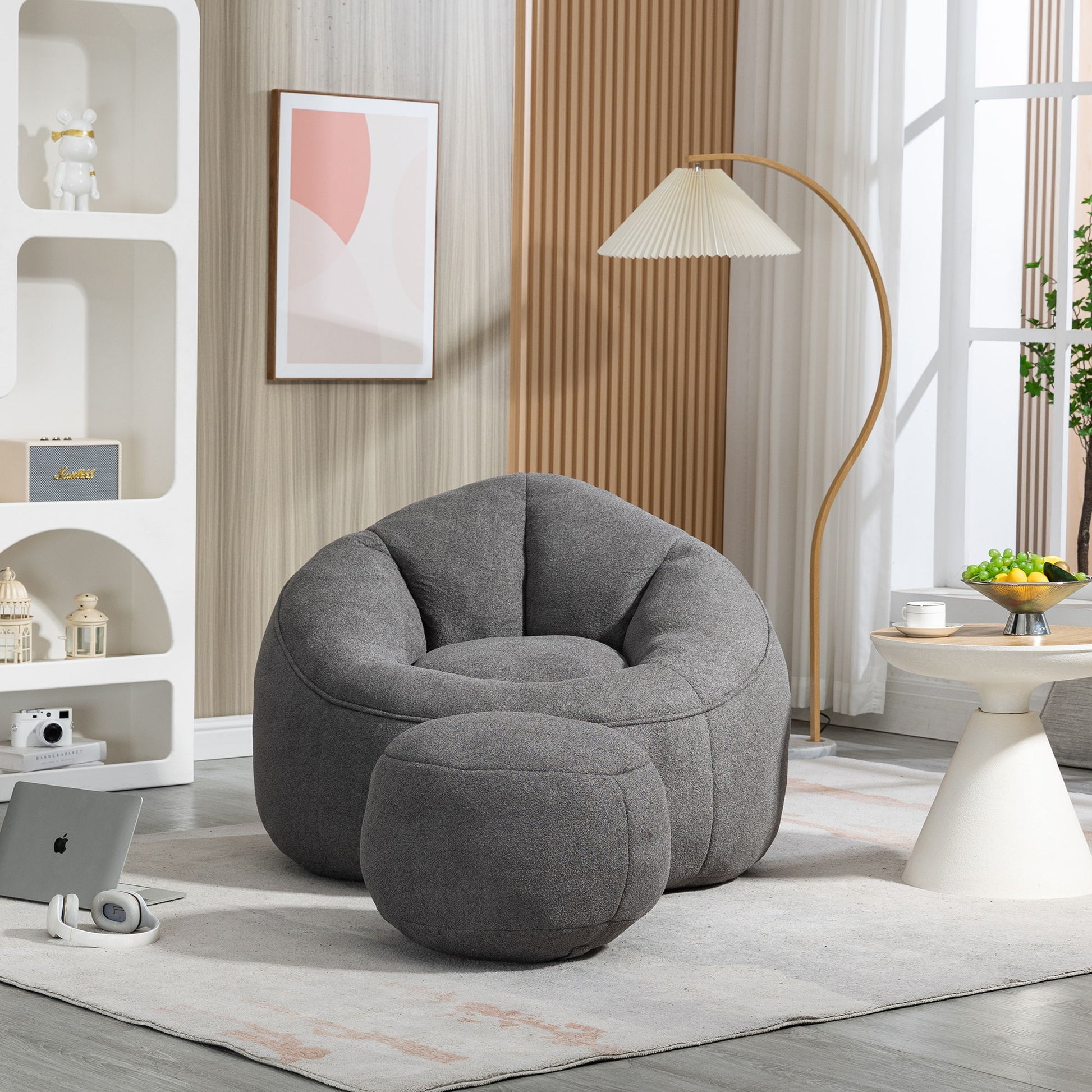 A modern living room with a gray Bedding Bean Bag Sofa Chair High Pressure Foam and matching footrest on a light rug, next to a side table with fruit bowl, white floor lamp, shelves with decor, and a painting on the wall.