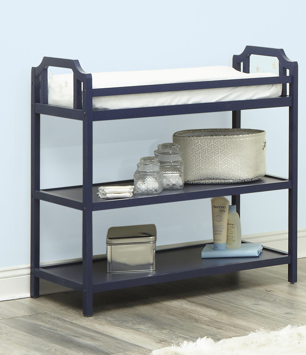 The Celeste Changing Table in navy blue features a white mattress on top, two open shelves with baskets, jars, wipes, lotion, and folded towels. It’s set against a light blue wall and rests on wood flooring.