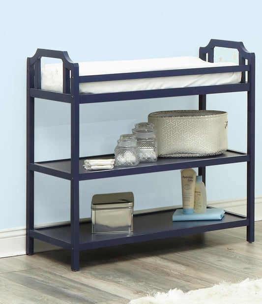 The Celeste Changing Table in navy blue features a white mattress on top, two open shelves with baskets, jars, wipes, lotion, and folded towels. It’s set against a light blue wall and rests on wood flooring.