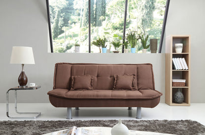 A modern living room featuring the Lionel Sofa Bed in brown with two matching cushions, a side table with a lamp, a wooden bookshelf filled with books and décor, and large windows offering views of greenery outside.