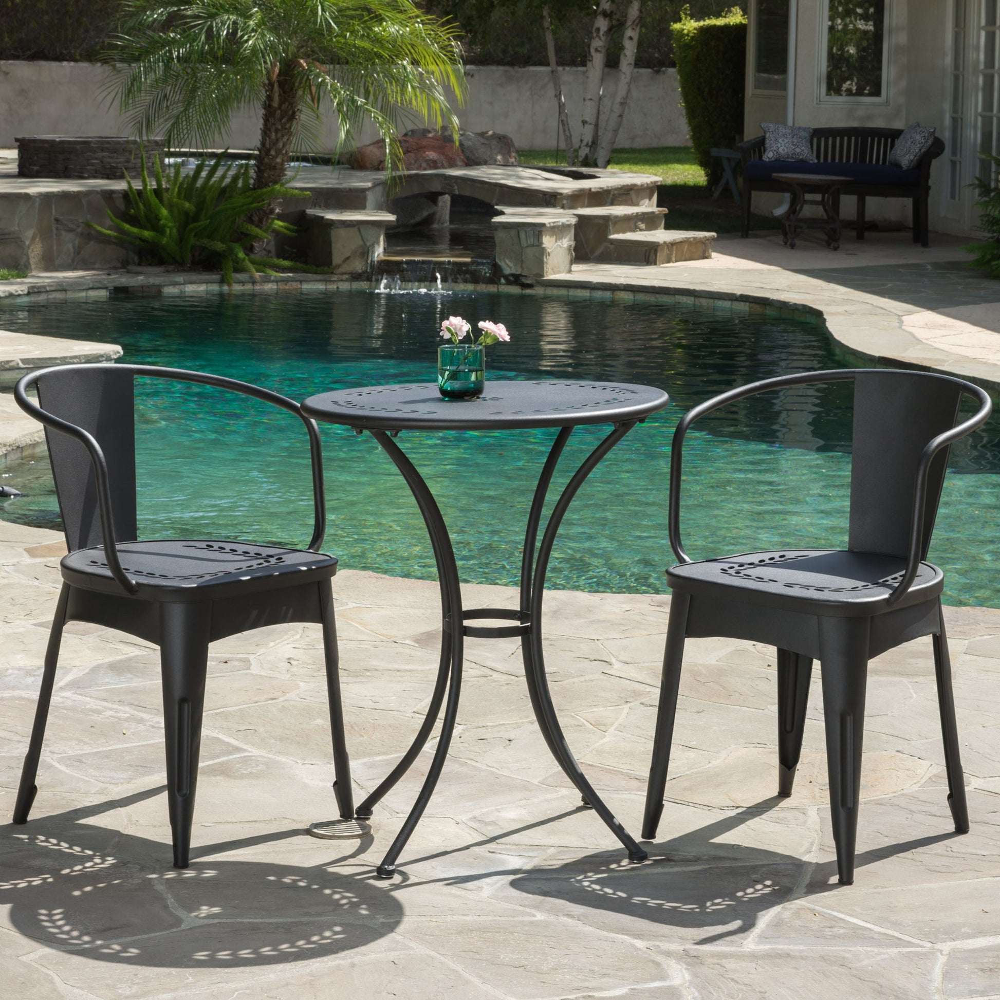 The Ambrosia 3 Piece Bistro Set with leaf motifs, featuring a small round table and two black metal chairs, sits on a stone patio by the pool. A small vase with pink flowers decorates the table, surrounded by greenery.
