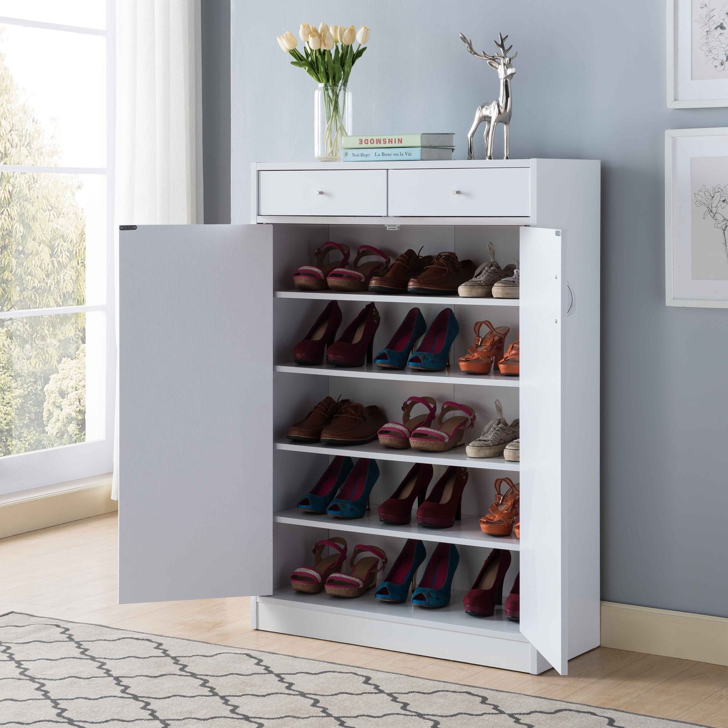 The Shoe Storage Cabinet with five shelves and two drawers fits 15 pairs of shoes. It stands in a bright, modern room with books, flowers in a vase, and a silver deer ornament displayed on top.